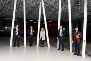 Cinq personnes se tiennent espacées les unes des autres dans le hall d'un immeuble moderne avec un plafond à motifs et de hauts piliers blancs.