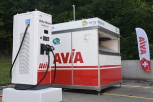 Borne de recharge pour véhicules électriques et unité de ravitaillement en hydrogène marquées « Avia » dans un parking, avec un arrière-plan boisé et la marque AVIA sur un drapeau.