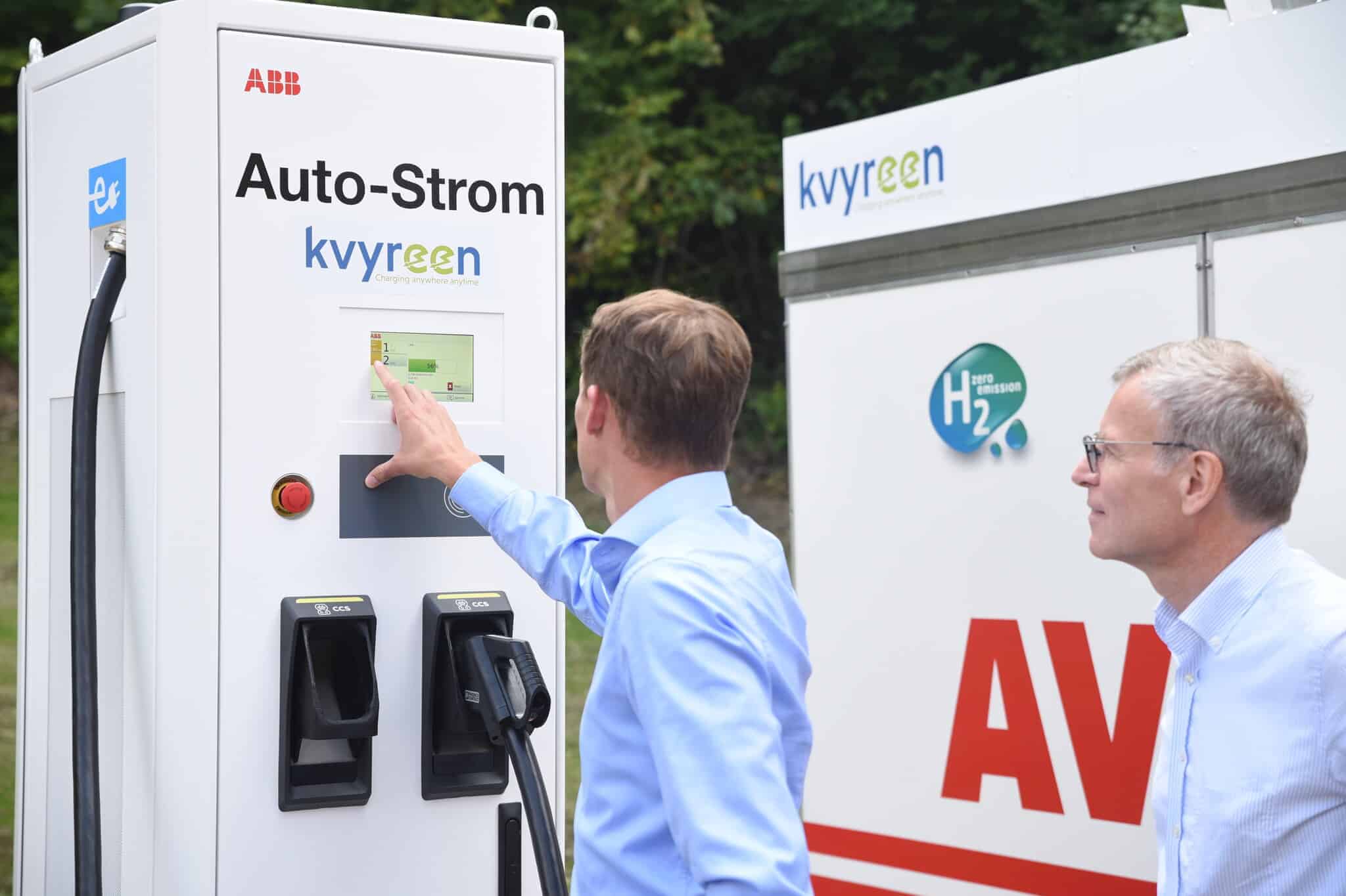 Deux hommes interagissent avec une station de recharge de véhicules électriques étiquetée «Auto-Strom» et «kvyreen» en plein air, avec une station d'hydrogène visible à l'arrière-plan.