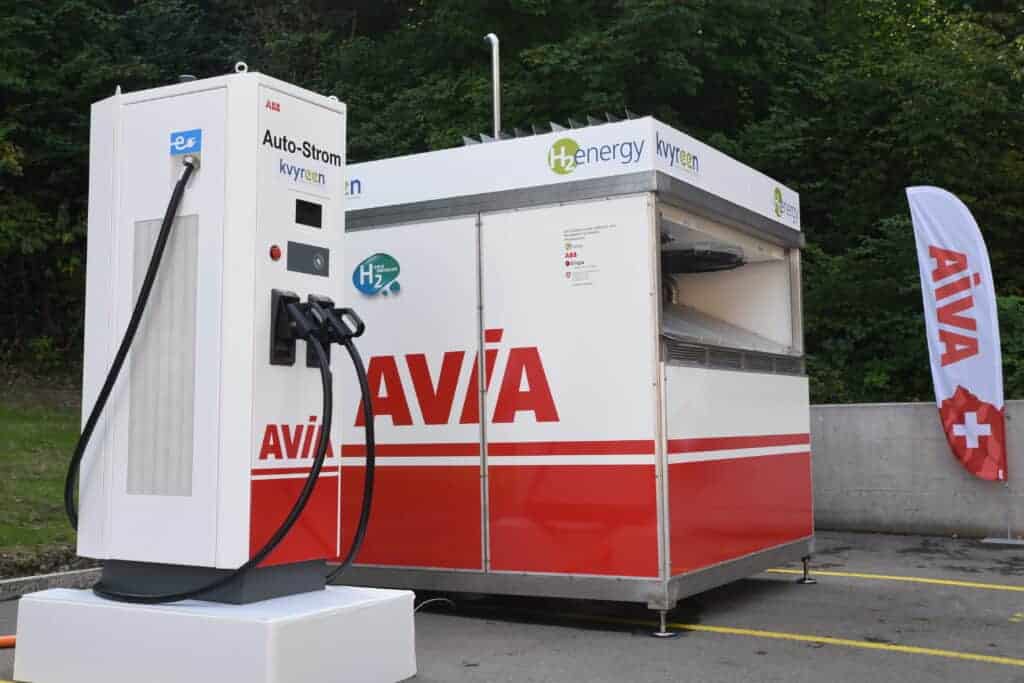 Ladestation für Elektrofahrzeuge und Wasserstofftankstelle mit der Aufschrift „AVIA“ auf einem Parkplatz, im Hintergrund Bäume und eine Betonmauer.