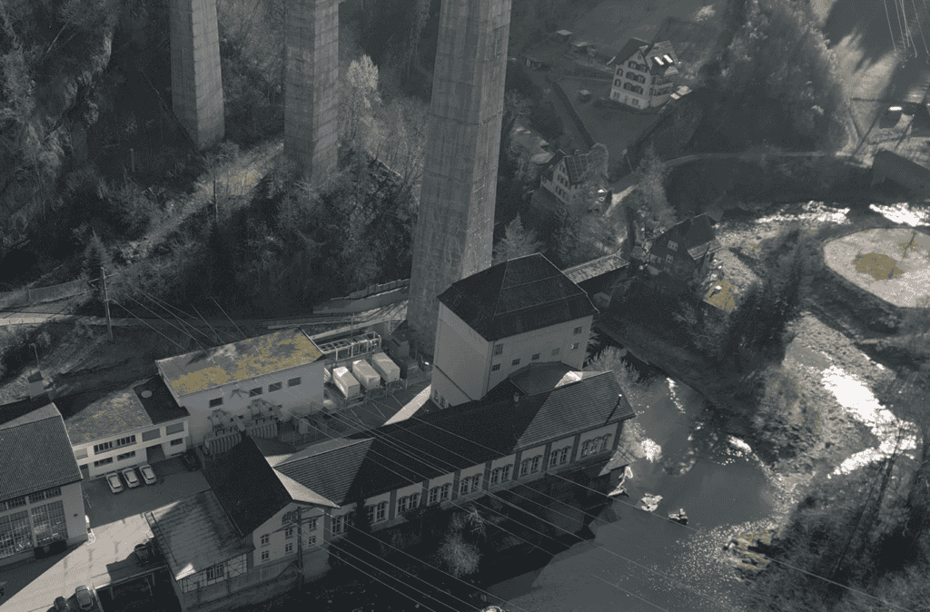 Vue aérienne d'une installation industrielle avec de grands bâtiments à côté d'une rivière, entourée de hauts piliers de pont en béton et de collines boisées.