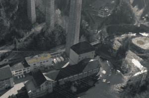 Vue aérienne d'une installation industrielle avec de grands bâtiments à côté d'une rivière, entourée de hauts piliers de pont en béton et de collines boisées.