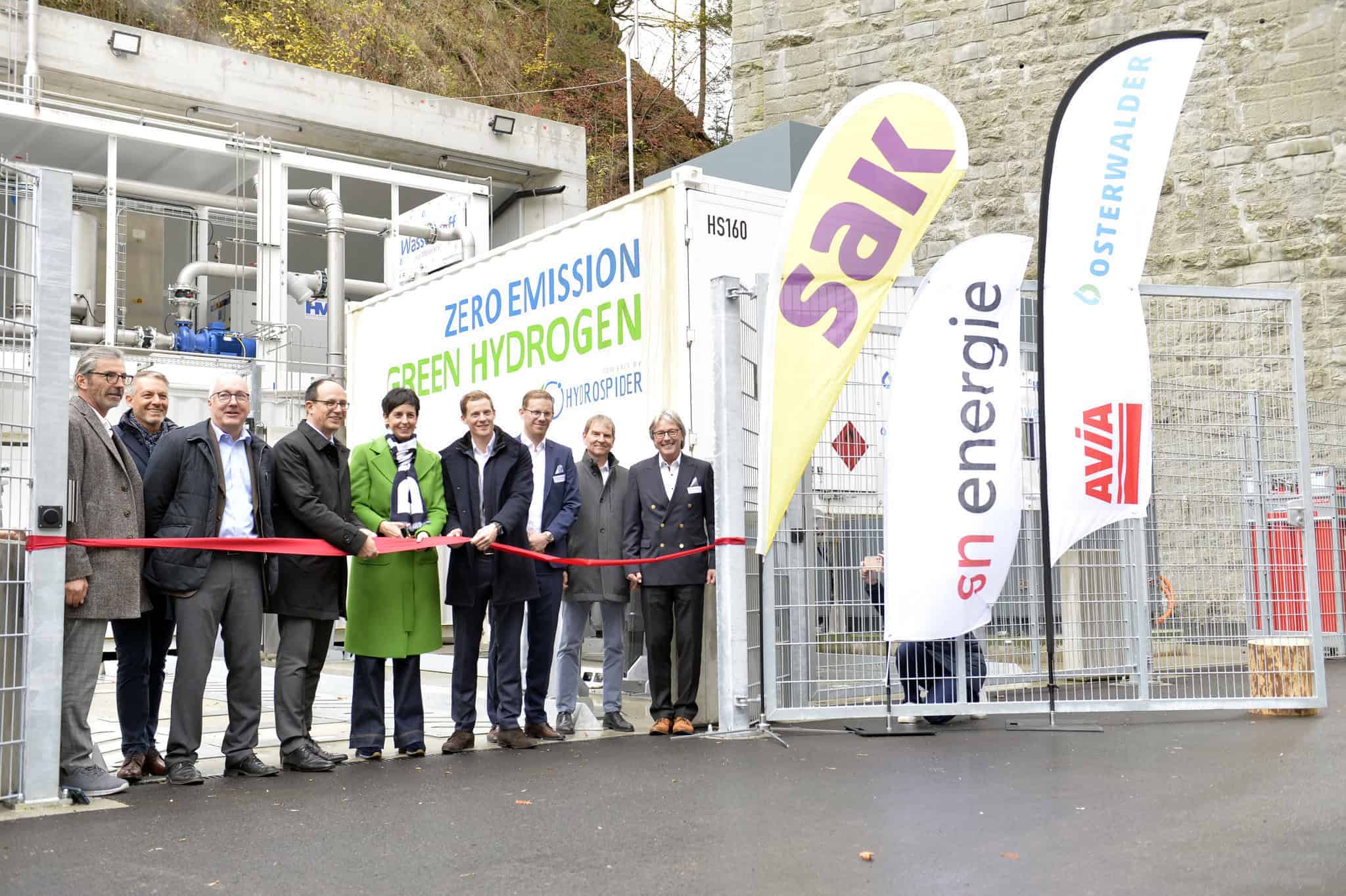 Un groupe de personnes se tient debout, coupant un ruban rouge lors de l'inauguration d'une installation de production d'hydrogène vert à zéro émission, avec des bannières de sponsors et des équipements industriels à l'arrière-plan.