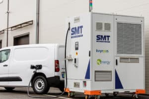Une camionnette électrique blanche est branchée sur une grande borne de recharge portable étiquetée « SMT » par Kvyreen et H2 Energy, positionnée à l'extérieur d'un bâtiment.