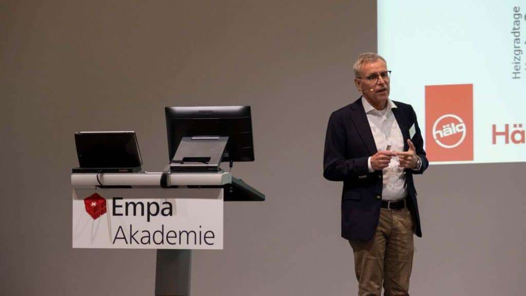 Un homme en costume se tient à côté d'un podium portant l'inscription « Empa Akademie » et fait une présentation avec un écran de projection derrière lui.