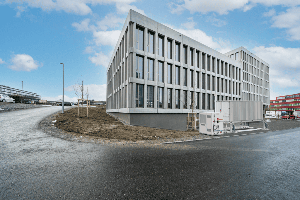 Un immeuble de bureaux moderne et rectangulaire, doté de hautes fenêtres verticales, se trouve à un angle ; une grande unité industrielle est installée à l'extérieur, près de la route.
