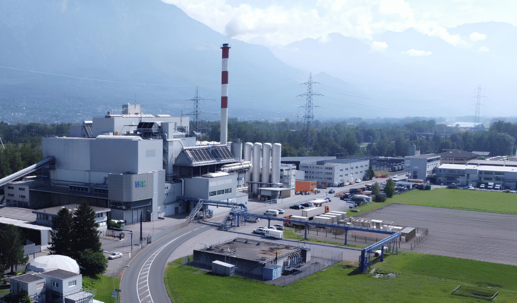 Une grande installation industrielle avec plusieurs bâtiments, une haute cheminée et plusieurs véhicules garés à l'extérieur, sur fond de montagnes et de verdure.