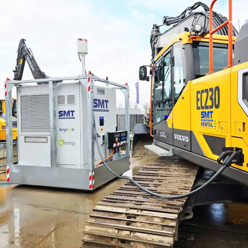 A large yellow Volvo excavator is connected to a white SMT charging station with hoses, parked on a concrete surface outdoors.