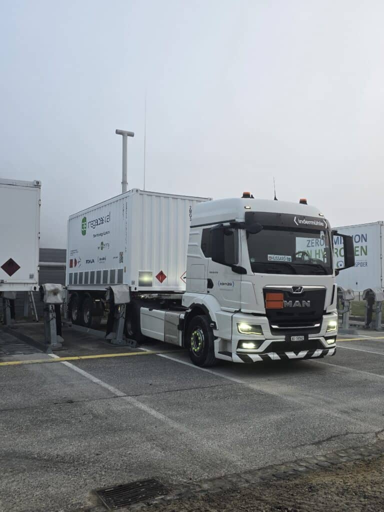 Ein weißer MAN-Sattelzug mit Wasserstoffaufkleber steht neben einer Lade- oder Tankstelle, an deren Anhänger Ladebehälter befestigt sind.