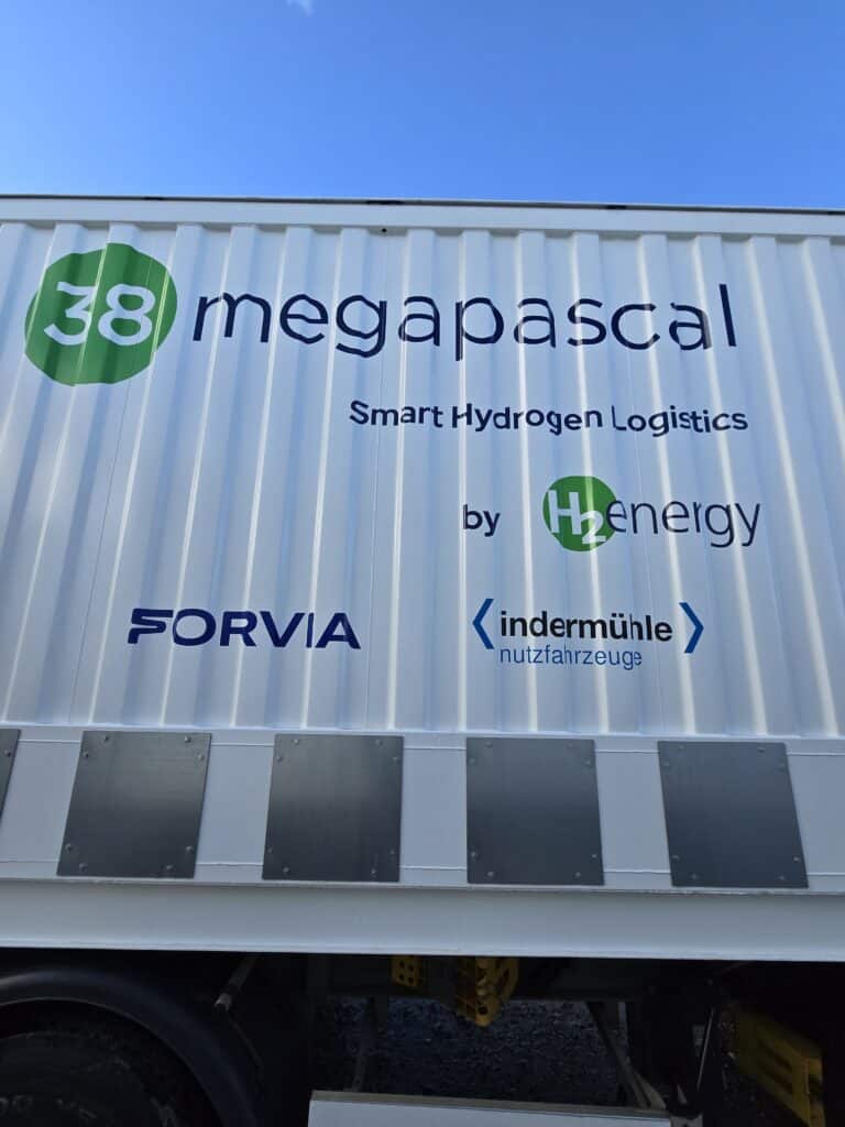 A white truck container displays the text "38 megapascal Smart Hydrogen Logistics by H2energy" along with Forvia and Indermühle logos against a clear blue sky.