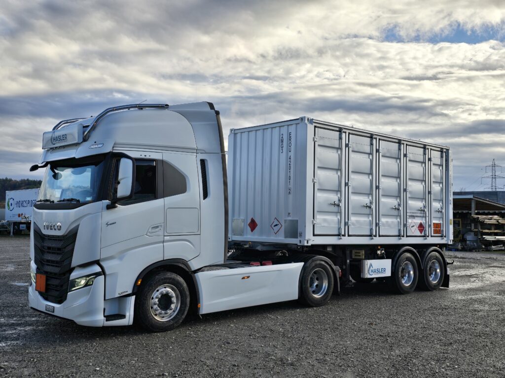 Ein weißer Iveco-Sattelzug ist auf einem Parkplatz geparkt, an dem ein Containeranhänger mit Gefahrgutwarnschildern befestigt ist, und der Himmel ist bewölkt.