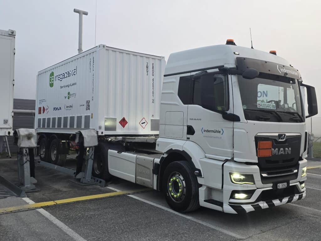 A white MAN truck with a large container on its trailer is parked at a facility checkpoint. The container displays various company logos and safety signs.