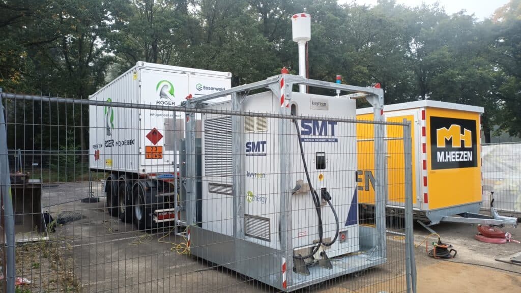 Fenced-in area with industrial equipment including a container labeled "SMT" and a white truck trailer with green logos; surrounded by trees.