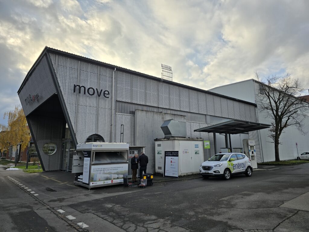 A modern industrial building with "move" on the facade, a white car labeled "enerv," and two people standing near charging or energy equipment outside.