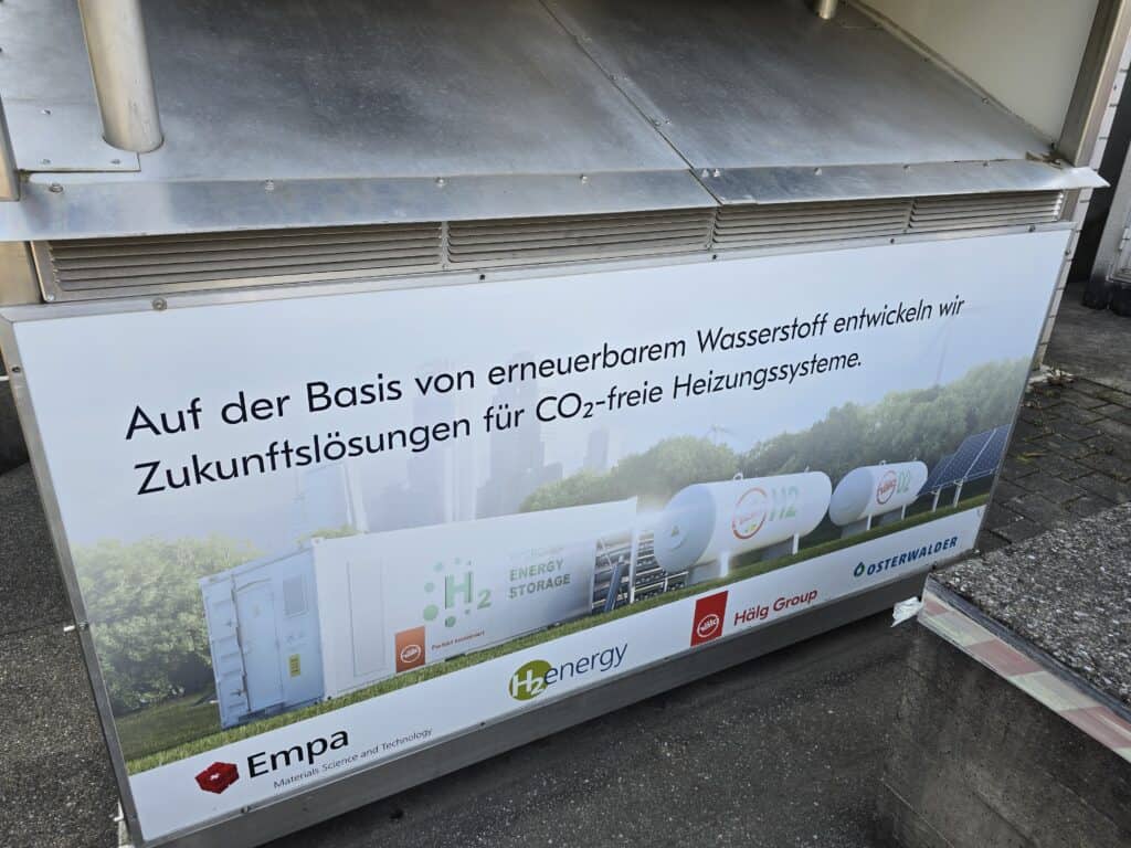 A sign in German promoting CO2-free heating systems using renewable hydrogen, featuring images of hydrogen storage containers, solar panels, and logos of associated organizations.