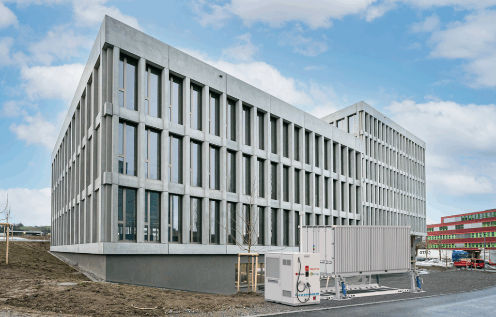 Modern office building with large rectangular windows; a white industrial HVAC unit is installed outside on a paved area in front of the building.